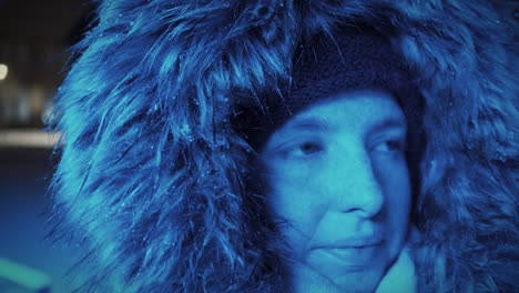 Young-woman-with-a-snow-covered-fur-hood-looking-around-in-front-of-a-blue-light-in-a-wintry-landscape-at-night---Close-up