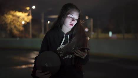 Teenage-girl-using-phone-holding-basketball-at-outdoor-court-at-night-in-city