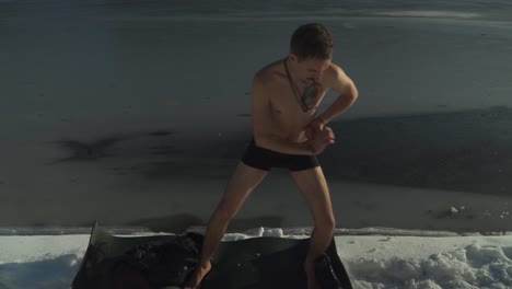Man-in-shorts-standing-on-black-yoga-mat-on-snow-covered-shoreline-of-frozen-mountain-lake-exercising-and-preparing-to-swim-in-ice-cold-water