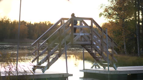 Young-hiking-boy-walking-on-wooden-bridge-over-beautiful-lake,-beautiful-autumn-scenery-at-sunset