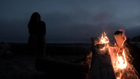 Girl-sitting-on-a-log-and-warming-up-next-to-camp-fire-at-night