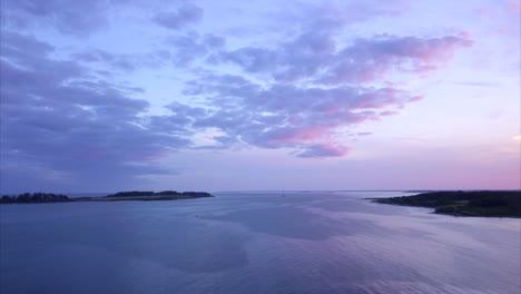 Aerial-shot-flying-out-over-a-calm-Atlantic-Ocean-harbor-during-a-colorful-soft-pink-and-purple-sunset-off-of-Kettle-Cove-Beach,-Maine.