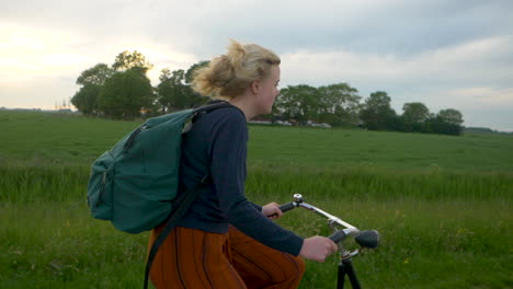 Young-blonde-girl-with-a-green-backpack-riding-along-a-green-field-with-some-trees-in-the-distance