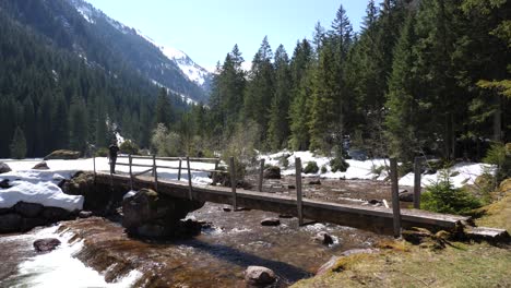 Still-shot-of-a-young-man-walking-over-a-wooden-bridge-on-a-hiking-route-in-the-Swiss-alps-over-a-calm-river-in-slow-motion