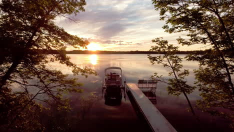 Flying-toward-docked-pontoon-boat-at-sunset-on-reflective-lake