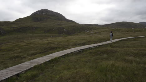 Girl-walking,-hiking-in-Connemara-National-Park-on-a-wooden-planked-trail-with-diamond-hill-in-the-background,-Ireland-4K