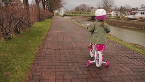 An-adorable-little-girl-in-pink-practicing-how-to-ride-a-bicycle-by-the-canal-in-spring-after-the-rain---slow-motion