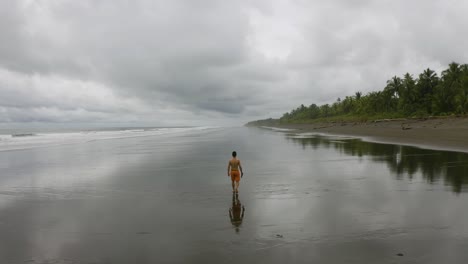 Aerial-follows-a-young-man-from-behind,-walking-at-an-empty-beach-in-Nuqui,-Colombia