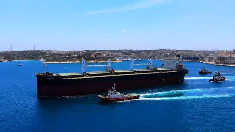 Dolly-out-aerial-shot-from-an-enormous-cargo-ship-leaving-the-harbour-of-Valletta,-Malta-with-the-help-of-three-tugboats