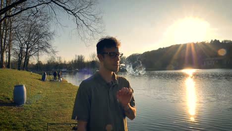 Man-standing-beside-a-lake-smoking-as-the-sun-sets-in-the-distance