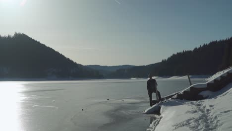 Ein-Mann-Steht-Am-Ufer-Des-Zugefrorenen-Sees-In-Siebenbürgen,-Rumänien-Und-Hält-An-Einem-Hellen-Wintertag-Ein-Stück-Holz-In-Der-Hand---Totale