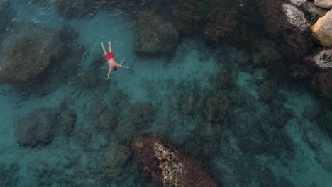 Young-Caucasian-man-swims-in-gorgeous-clear-turquoise-water-by-large-rocks,-circle-aerial-shot