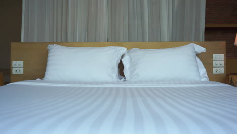 Hotel-bed-with-white-striped-bedspread-and-pillows-and-wind-blowing-curtains