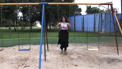Young-woman-with-long-skirt-and-colorful-shirt-swinging-and-laughing-feeling-free-and-happy-at-a-park-viewed-from-the-front-with-subtle-camera-movement