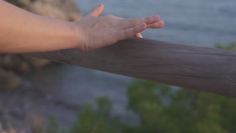 The-hand-of-a-woman-caresses-and-feels-the-touch-of-the-wood-of-a-trunk-shaped-like-a-handrail-that-is-located-on-the-coast,-in-the-background-you-can-see-the-blue-sea-at-sunset