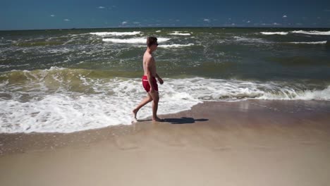 Junger-Mann-In-Roten-Shorts-Geht-An-Einem-Strand-Am-Meer-Spazieren