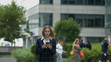 In-a-clean-urban-setting,-a-young-women-walks-towards-the-camera-paying-extreme-attention-to-her-cell-phone