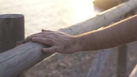 The-hand-of-a-man-caresses-and-sinete-the-touch-of-a-trunk-as-a-railing-during-a-sunset-on-the-coast,-in-the-golden-background-of-the-image-you-can-see-the-sea