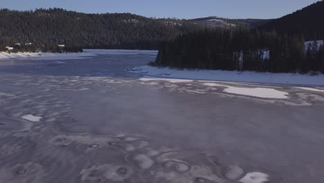 Ascending-aerial-view-from-drone-over-frozen-wilderness-lake-in-winter-with-ice-and-snow-on-surface-and-terrain