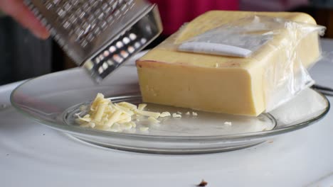 Close-up-on-male-hands-picking-up-sliced-block-of-yellow-cheddar---grating-it-into-a-pile-on-small-glass-plate-in-home-kitchen
