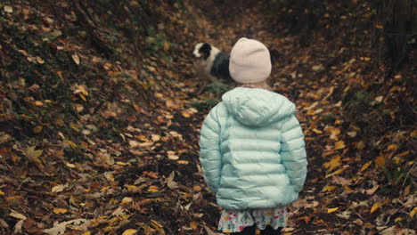 Young-girl-in-winter-coat-follows-dog-on-autumn-path-in-woods,-slow-mo