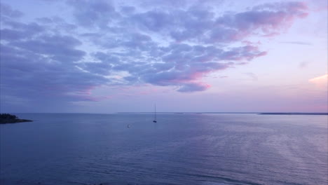 Luftaufnahme,-Die-Mit-Einem-Einzelnen-Segelboot-Während-Eines-Dramatischen-Rosa-Und-Violetten-Sonnenuntergangs-Vor-Der-Küste-Von-Maine-über-Einen-Ruhigen-Hafen-Fliegt