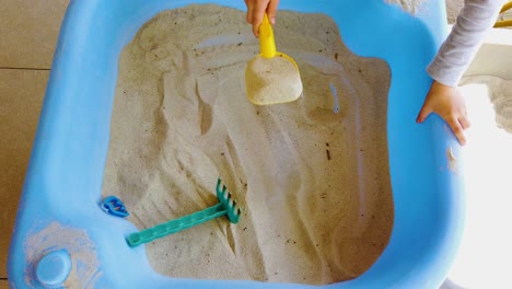 Child-playing-in-blue-sandpit-scooping-up-sand-with-plastic-shovel