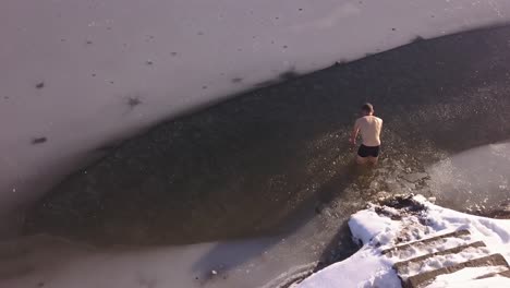 Aerial-view-of-young-man-entering-frozen-lake-in-winter-through-broken-ice