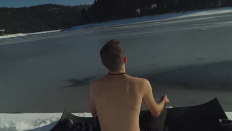 View-from-behind-man-without-shirt-meditating-on-black-yoga-mat-on-snow-covered-shoreline-of-frozen-alpine-lake