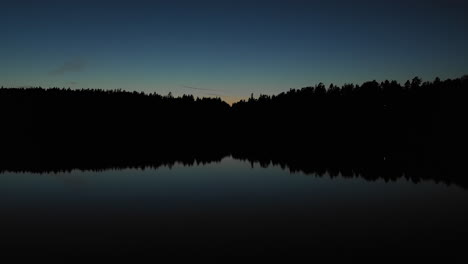Aerial,-drone-shot,-close-to-the-water,-above-a-lake-and-towards-dark-forest,-colorful-dusk,-in-Sweden