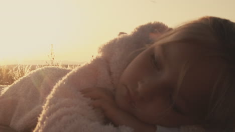 Pan-around-little-girl-snuggled-in-warm-fuzzy-jacket-on-picnic-table-as-setting-sun-glows-behind-her
