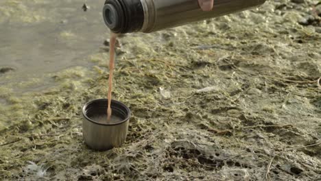 Pouring-hot-drink-from-flask-by-lake-side-shore