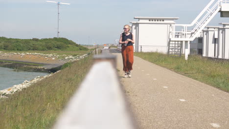 A-young-blonde-female-wearing-sunglasses-orange-pants-and-blacktop-walking-along-a-footpath-holding-a-bottle-of-water