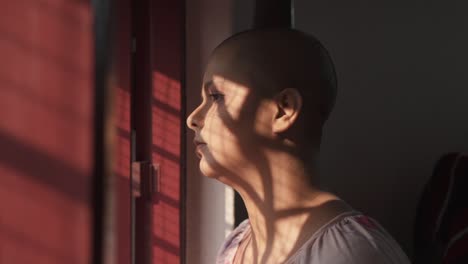 Sad-and-lonely-bald-Indian-girl-standing-near-window-and-looking-outside-from-home-due-to-Pandemic-covid-19-lockdown