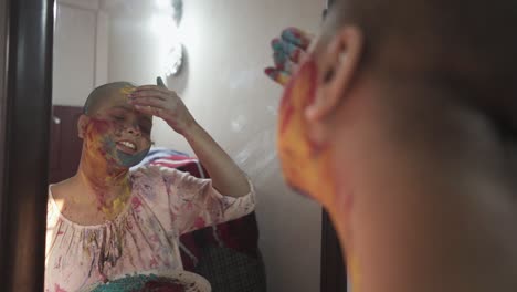 Hopeful-cancer-patient-bald-girl-applies-colored-powder-on-cheeks-at-home-in-front-of-mirror-on-Holi,-Indian-festival-of-colors