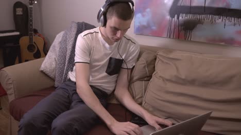 Young-Man-Sitting-On-The-Sofa-And-Working-With-His-Laptop-While-Listening-To-Music-At-Home