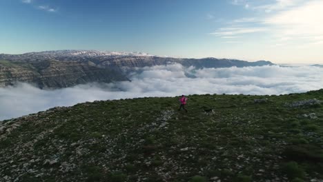 Man-runs-with-dog-on-rocky-mountain-top-with-view-of-clouds-in-valley,-aerial