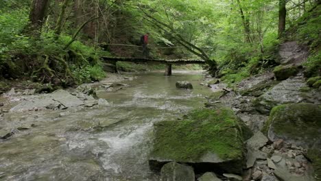 Man-walking-over-a-wooden-bridge-in-the-forest-as-the-river-stream-runs-in-the-foreground-and-under-the-bridge