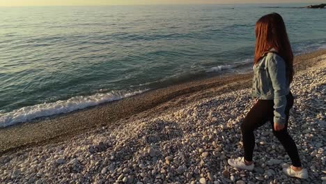 Caucasian-woman-walking-on-rocky-beach-towards-ocean-and-sunshine,-Kafarabida,-Lebanon,-spinning-aerial