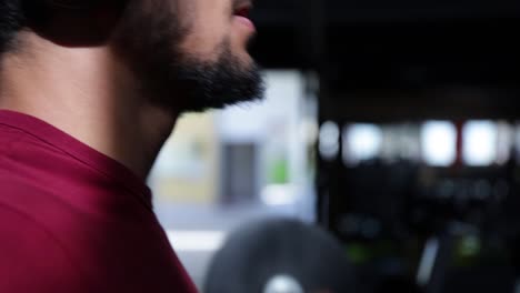 Young-Caucasian-bearded-man-wearing-hat-and-headphones-doing-biceps-workout-with-barbell-in-gym,-close-up-profile