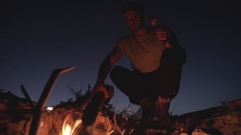 Caucasian-man-moving-dead-wood-log-in-firepit-on-bright-clear-evening-sky-with-half-moon-in-distance,-low-angle-close-up-pan