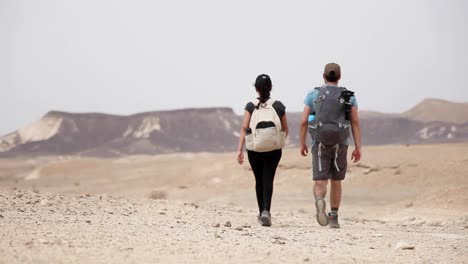Adventurer-couple-walking-through-the-valley,-mountains-at-the-background,-Israel-desert,-cloudy-day,-static-shot