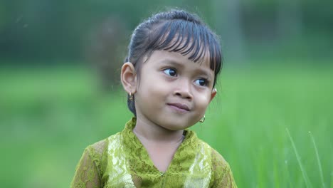A-cute-little-girl-dressed-in-a-traditional-Balinese-dress-is-standing-between-rice-fields-on-her-family's-property,-smiling-directly-to-the-camera