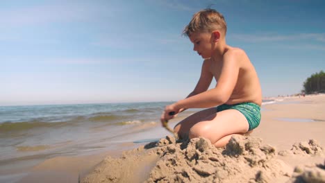 Young-boy-in-swimwear-plays-at-the-beach-and-digs-a-path-into-the-sand-with-shovel