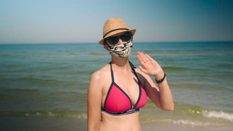 Junge-Bikinifrau-Am-Strand-Mit-Corona-Gesichtsmaske-Winkt-In-Die-Kamera