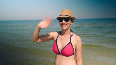 Junge-Bikini-Frau-Zieht-Korona-Gesichtsmaske-Am-Strand-Aus,-Winkt-Und-Lächelt