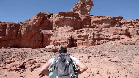 Adventurer-with-backpack-walking-through-arid-desert,-stream-valley,-mountains-at-the-background,-blue-sky,-wide-angle-shot,-traveling