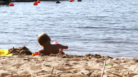 Small-blonde-boy-plays-in-sand-by-water-line-on-hot-day-on-sunny-beach