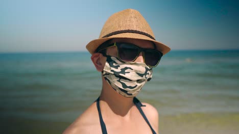 Frau-Trägt-Corona-Gesichtsmaske-Mit-Sonnenbrille-Und-Hut-Am-Strand-Neben-Dem-Meer