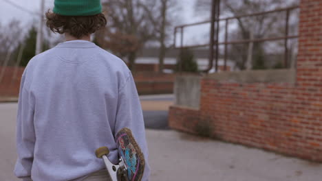 Teen-boy-with-a-skateboard-walks-away-and-towards-a-back-alley-parking-lot
